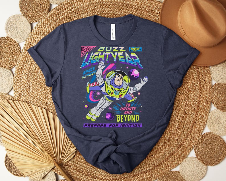 May include: Navy blue t-shirt featuring a colorful graphic of Buzz Lightyear in a space scene. The design includes the text "Buzz Lightyear" and "To Infinity and Beyond." The shirt has a crew neck and short sleeves.