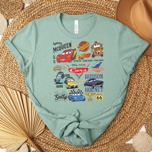 Puede incluir: Una camiseta verde claro con un gr&aacute;fico colorido de personajes de la pel&iacute;cula animada "Cars". El dise&ntilde;o incluye a Rayo McQueen, Mate, Sally y otros personajes, junto con el texto "Cars" y "Route 66".