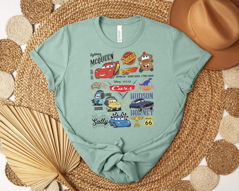 Puede incluir: Una camiseta verde claro con un gr&aacute;fico colorido de personajes de la pel&iacute;cula animada "Cars". El dise&ntilde;o incluye a Rayo McQueen, Mate, Sally y otros personajes, junto con el texto "Cars" y "Route 66".