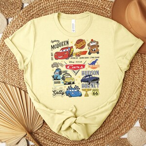 Puede incluir: Una camiseta de color amarillo claro con un dise&ntilde;o gr&aacute;fico colorido de personajes y logotipos de la pel&iacute;cula "Cars". El dise&ntilde;o incluye a Rayo McQueen, Mate y otros personajes, junto con el texto "Cars" y "Route 66".