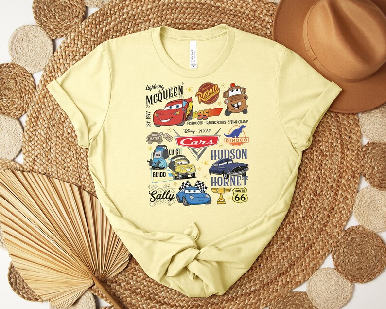 Puede incluir: Una camiseta de color amarillo claro con un dise&ntilde;o gr&aacute;fico colorido de personajes y logotipos de la pel&iacute;cula "Cars". El dise&ntilde;o incluye a Rayo McQueen, Mate y otros personajes, junto con el texto "Cars" y "Route 66".