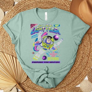 May include: A light green t-shirt featuring a graphic of Buzz Lightyear from Toy Story. The design includes the words "Buzz Lightyear", "Space Ranger", and "To Infinity and Beyond". The shirt has a crew neck and short sleeves.