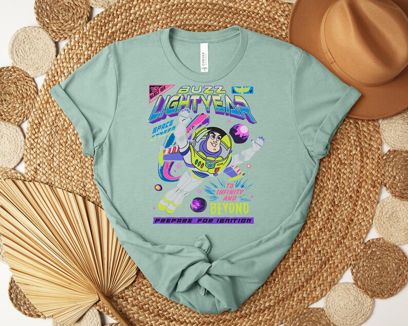 May include: A light green t-shirt featuring a graphic of Buzz Lightyear from Toy Story. The design includes the words "Buzz Lightyear", "Space Ranger", and "To Infinity and Beyond". The shirt has a crew neck and short sleeves.