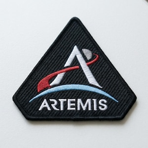 May include: Black embroidered patch with the Artemis logo. The logo features a white stylized letter "A" with a red and white swoosh, a blue arc, and the word "ARTEMIS" in white. The patch has a black border and a triangular shape.