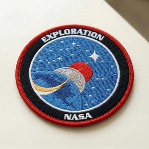 May include: Embroidered patch with a space exploration theme. The patch features a blue background with stars, a red and silver planet, and the text "EXPLORATION" and "NASA". The patch has a black border with a red outer edge.