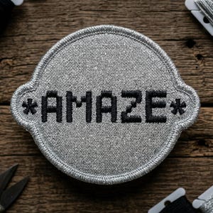 May include: A silver embroidered patch with the word "AMAZE" in black block letters. The patch has a decorative border and asterisks on each side of the word. The patch is on a wooden surface, with spools of thread and scissors in the background.