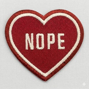 May include: A heart-shaped patch in a deep red color with a white border. The word "NOPE" is embroidered in white block letters across the center of the patch. The patch is likely made of fabric.