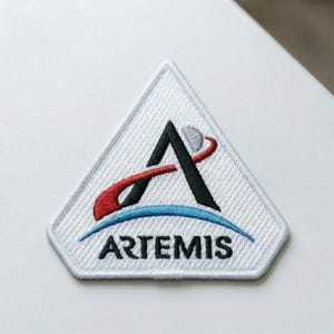 May include: Embroidered patch featuring the Artemis program logo. The design includes a black stylized letter "A" with a red and white arc, a blue arc, and the word "ARTEMIS" in black text. The patch has a white background and a geometric shape.