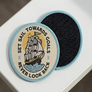 May include: Two round patches with a light blue border. One patch features a sailing ship illustration with the text "SET SAIL TOWARDS GOALS NEVER LOOK BACK". The other patch has a black fabric backing.