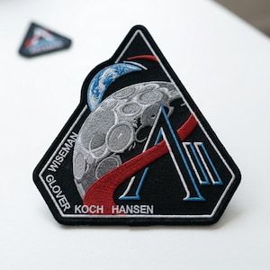 May include: Embroidered black patch with a triangular shape, featuring a detailed depiction of the moon, Earth, and a stylized letter "A". The patch includes the names "WISEMAN", "GLOVER", "KOCH", and "HANSEN". A smaller patch is visible in the background.