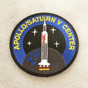May include: Embroidered patch featuring the Apollo/Saturn V Center logo. The patch is circular with a blue border and yellow text. The design includes a rocket, stars, and a depiction of the moon. The word "USA" is visible on the rocket.