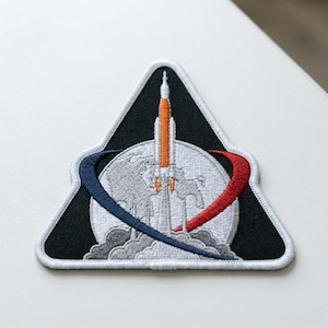 May include: Embroidered patch featuring a rocket launching from Earth, with a black triangle border and a white outline. The design includes a blue and red arc, and a detailed depiction of the Earth.