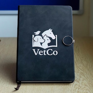 Puede incluir: Cuaderno negro con un logotipo blanco que presenta un perro, un gato, un pájaro y un caballo, con el texto "VetCo". El cuaderno tiene un cierre magnético y una cinta de marcador. El cuaderno está sobre una superficie de madera.