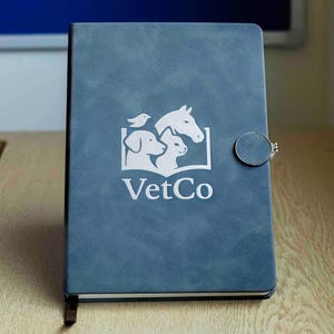 May include: A blue notebook with a silver clasp. The cover features a white logo with a dog, cat, bird, and horse, along with the text "VetCo". The notebook is closed and has a bookmark.