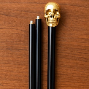 May include: A decorative cane with a gold skull handle and a black shaft. The cane is shown with two additional black shaft segments. The cane is displayed on a brown wooden surface.