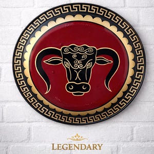 May include: A round, decorative plate with a red center featuring a black bull's head design. The plate has a gold border and a black outer rim with a Greek key pattern. The word "LEGENDARY" is printed below.