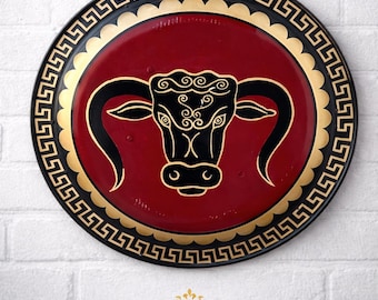 May include: A round, decorative plate with a red center featuring a black bull's head design. The plate has a gold border and a black outer rim with a Greek key pattern. The word "LEGENDARY" is printed below.