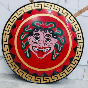 May include: A round, decorative shield featuring a Medusa design. The shield has a red center with a Medusa face and green snakes. The outer rim is black with a gold Greek key pattern.