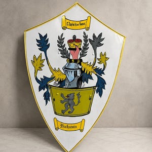May include: A white heater shield with a gold border featuring a coat of arms. The design includes a yellow base with a grey lion, a knight's helmet, a hand holding a crown, and scrolls with the text Clario hinc honos and Buchanan.
