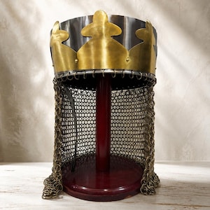 May include: A metal crown helmet featuring a polished steel cylindrical band topped with a brass fleur-de-lis rim. A brass chainmail aventail hangs from the base of the crown, secured by small metal rings. The helmet is displayed on a dark red stand.