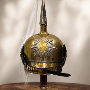May include: A brass and silver military helmet with a decorative crest and a white plume. The helmet features a detailed emblem and a black chin strap. The word "LEGENDARY" is displayed at the base.