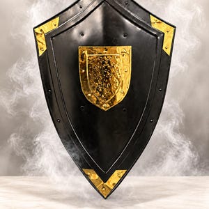 May include: A black heater shield featuring a central gold-colored hammered metal plate and gold-colored corner accents secured with rivets. The word LEGENDARY is printed in gold at the bottom center of the shield's surface.