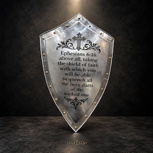 May include: A silver-toned metal shield with a pointed base and rounded top, featuring gold-colored rivets along the perimeter. The center displays a black engraved cross, decorative scrollwork, and the text Ephesians 6:16 above all, taking the shield of faith with which you will be able to quench all the fiery darts of the wicked one.