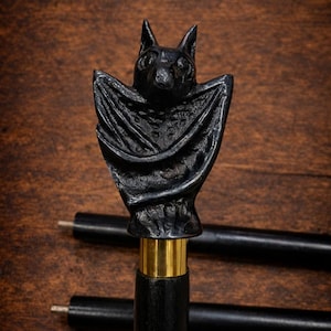 May include: A black walking cane with a bat-shaped handle. The bat is carved with detailed wings and a textured body. The handle is connected to the cane with a gold-colored band. The cane is black and has a smooth finish.