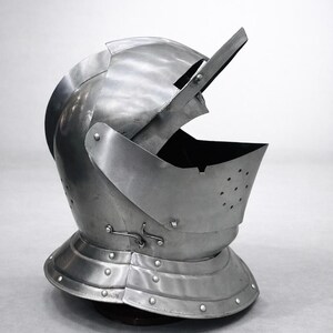 May include: A silver-colored medieval helmet with an open visor. The helmet has a rounded top, a neck guard, and is riveted together. The visor has small holes for vision and ventilation. The helmet is designed to protect the head and face.
