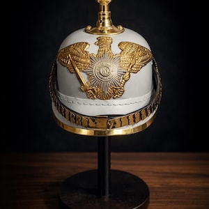 May include: A white and gold military helmet with an ornate design, featuring a gold eagle emblem and a decorative band. The helmet is displayed on a black stand, with the word "LEGENDARY" at the base.