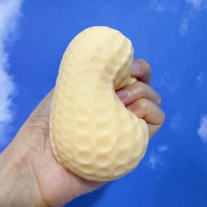 Puede incluir: Una bola antiestrés texturizada de color amarillo claro con forma de cacahuete se sostiene en una mano sobre un fondo de cielo azul. La bola antiestrés tiene una superficie con hoyuelos. La mano es parcialmente visible, apretando la bola.