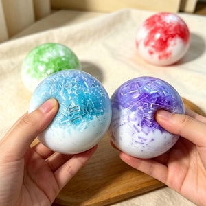 May include: A collection of colorful, spherical stress balls. One is held in a hand, showing a blue and white design. Other balls feature green, red, and purple patterns. The balls have a textured, crystalline appearance.