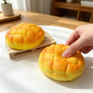 May include: Two yellow and orange, textured, round objects resembling bread rolls. One sits on a small wooden pallet, while a hand gently touches the other. The objects have a grid-like pattern on top.