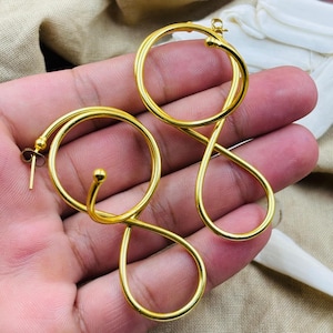 Puede incluir: Pendientes dorados con un diseño abstracto único. Uno presenta un aro circular conectado a una forma de lágrima, mientras que el otro tiene un diseño en forma de ocho. Los pendientes se muestran sobre un fondo neutro.