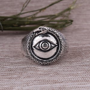 Op de afbeelding: Een zilverkleurige ring met een oogontwerp in het midden, omcirkeld door een slang die zijn staart opeet. De ring heeft een getextureerde band en een gedetailleerd, antiek uiterlijk. Het oog is een prominent middelpunt.