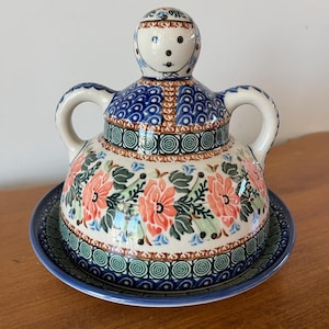 May include: A ceramic butter dish with a lid, featuring a hand-painted floral design in shades of red, blue, and green. The dish has a round base and a lid shaped like a person with handles on the sides. The dish sits on a wooden surface.