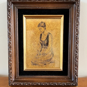 May include: A framed artwork featuring a sketch of a person kneeling, set against a gold background. The artwork is encased in a dark brown, ornate wooden frame with a black velvet matting.