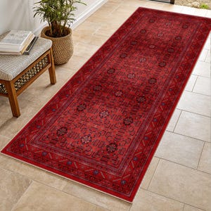 May include: A rectangular red rug with an intricate geometric pattern. The rug features a central field of repeating shapes and floral motifs, framed by a detailed border. The rug is designed for a hallway or entryway.