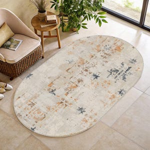 May include: An oval area rug with a distressed design. The rug features a cream-colored background with accents of orange, brown, and navy blue. The rug is placed on a tiled floor, and a chair and side table are visible in the background.