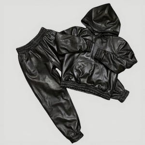 Puede incluir: Un conjunto de ch&aacute;ndal de cuero negro, que incluye una sudadera con capucha y pantalones a juego. La sudadera tiene un bolsillo delantero y una capucha con cord&oacute;n. Los pantalones tienen cintura y pu&ntilde;os el&aacute;sticos. El conjunto es de polipiel brillante.
