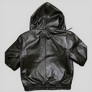 Puede incluir: Chaqueta con capucha de cuero negro con mangas cortas. La chaqueta presenta una capucha con cord&oacute;n, un dobladillo acanalado y un dise&ntilde;o cl&aacute;sico de chaqueta bomber. El cuero tiene un aspecto ligeramente texturizado.