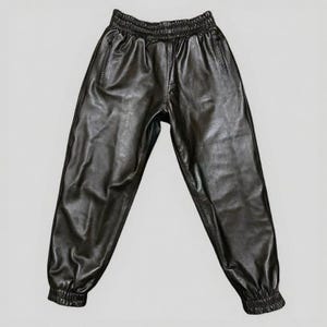 Puede incluir: Pantalones deportivos de cuero negro con cintura y pu&ntilde;os el&aacute;sticos. Los pantalones tienen un corte relajado con un ligero brillo, con dos bolsillos laterales y una cremallera. El material parece suave y flexible.