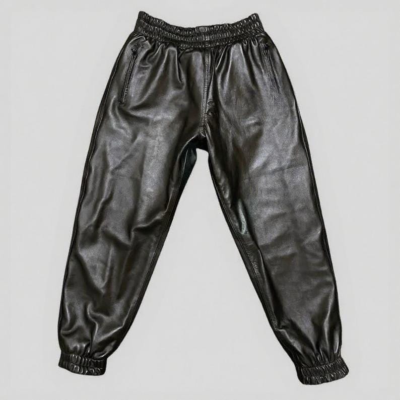 Puede incluir: Pantalones deportivos de cuero negro con cintura y pu&ntilde;os el&aacute;sticos. Los pantalones tienen un corte relajado con un ligero brillo, con dos bolsillos laterales y una cremallera. El material parece suave y flexible.