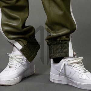 May include: Close-up of white sneakers with white laces and olive green leather pants with white side stripes. The pants have a zipper detail at the ankle and a ribbed cuff. The sneakers are on a gray background.