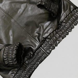 Puede incluir: Primer plano de una chaqueta de cuero negra. La chaqueta presenta una superficie brillante y texturizada con pu&ntilde;os y dobladillo el&aacute;sticos fruncidos. Las costuras son visibles y el dise&ntilde;o sugiere un estilo bomber o similar.