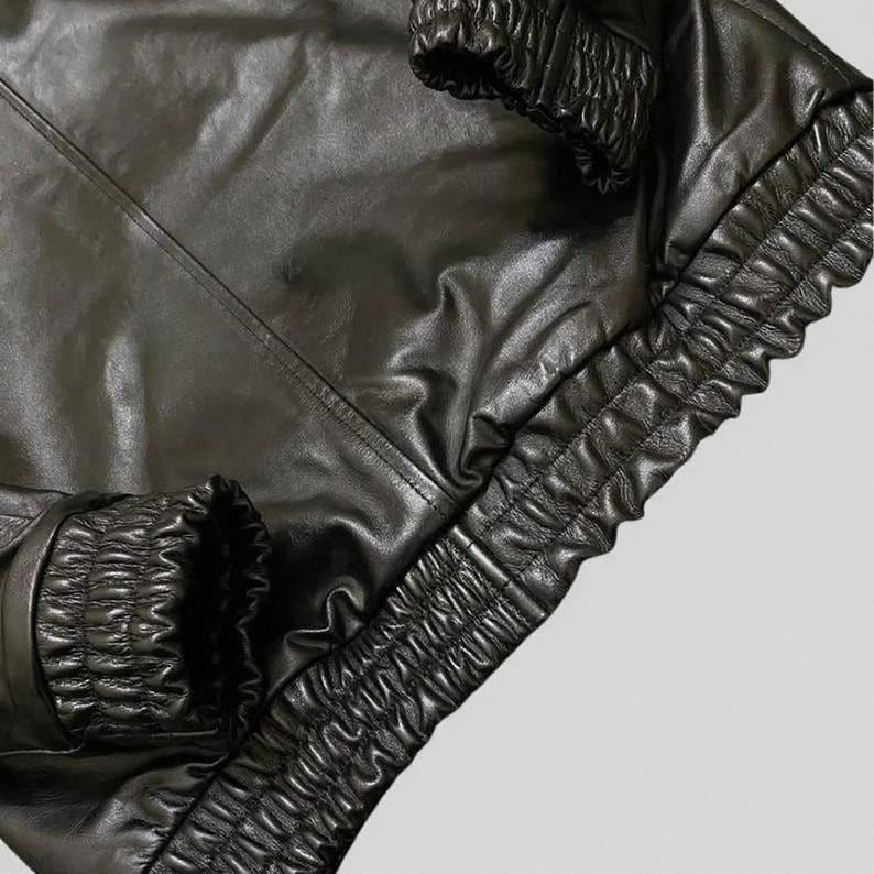 Puede incluir: Primer plano de una chaqueta de cuero negra. La chaqueta presenta una superficie brillante y texturizada con pu&ntilde;os y dobladillo el&aacute;sticos fruncidos. Las costuras son visibles y el dise&ntilde;o sugiere un estilo bomber o similar.
