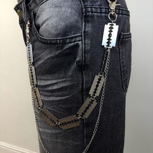 May include: Silver-colored chain belt featuring razor blade pendants. The belt is attached to the waistband of dark gray denim jeans with a silver clasp. The chain is made of small, linked metal pieces.
