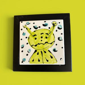 May include: A square ceramic tile framed in black, featuring a hand-painted lime green alien character with two antennae, three eyes, and a jagged mouth. The background is white with scattered black dots and small teal blue circular accents.