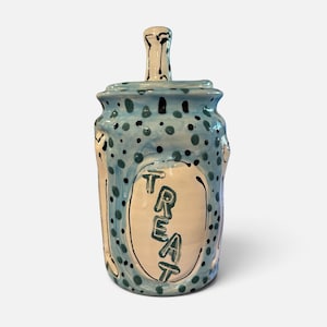 May include: A cylindrical ceramic jar with a lid featuring a vertical handle. The jar is light blue with dark blue and black polka dots. A central white oval panel on the front displays the word TREAT in dark blue, hand-painted capital letters.