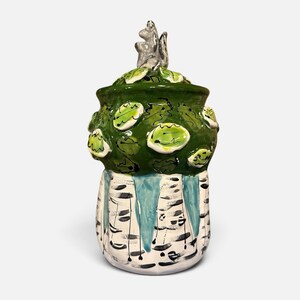 May include: A ceramic jar with a lid featuring a sculpted squirrel finial. The upper section is dark green with raised light green leaf motifs. The lower section is white with black horizontal markings and vertical light blue drip patterns.